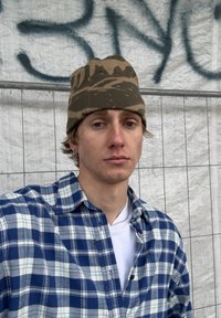 Personne portant une chemise à carreaux bleue et blanche sur un t-shirt blanc, ainsi qu'un bonnet marron camouflage. Porte des boucles d'oreilles percées et a les cheveux mi-longs.