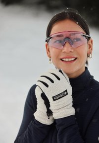 Gants en polaire blanche avec des accents noirs, portés par une personne, accompagnés de lunettes de ski teintées rose et d'un haut zippé sombre, sur fond enneigé.