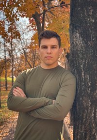 Μακρυμάνικο ελαιόχρωμο πουκάμισο, χαλαρή εφαρμογή, στέκεται μπροστά σε ένα δέντρο με πορτοκαλί φθινοπωρινά φύλλα στο φόντο. Φυσικός εξωτερικός φωτισμός.