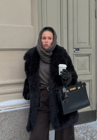 Mujer con abrigo de piel negro y capucha, sosteniendo una taza de café y un bolso negro con gafas de sol, de pie cerca de una puerta beige de un edificio en clima frío.