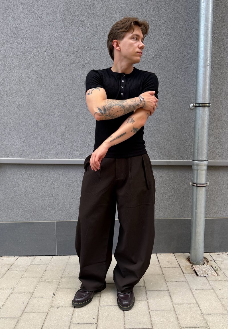 Jeune personne avec des tatouages sur les bras, portant une chemise noire et un pantalon large marron, debout sur un sol carrelé près d’un mur gris avec un tuyau métallique.