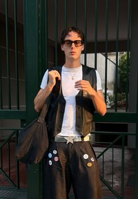 Veste noire en cuir portée sur un t-shirt blanc, pantalon noir ample avec plusieurs boutons, boucle de ceinture en forme d'étoile argentée, lunettes de soleil, debout devant des barres vertes.