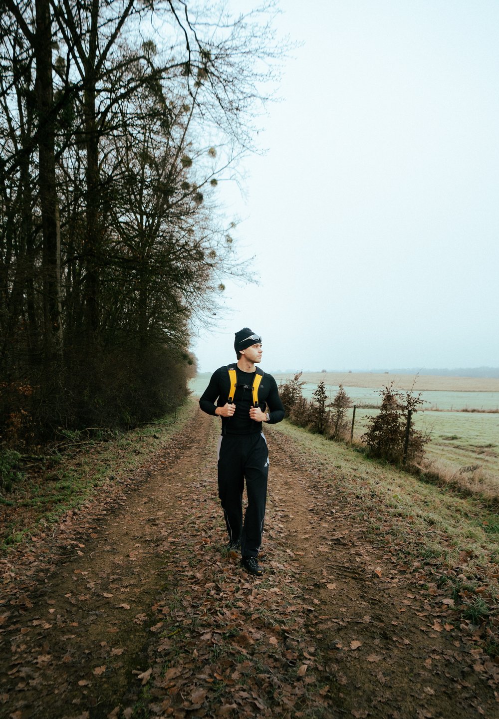 Egy fekete hosszú ujjú pólót és fekete nadrágot viselő személy sétál egy földúton, élénksárga hátizsákot és fekete sapkát viselve.