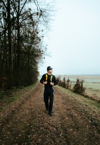 Egy fekete hosszú ujjú pólót és fekete nadrágot viselő személy sétál egy földúton, élénksárga hátizsákot és fekete sapkát viselve.