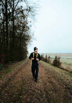Una persona con una camisa de manga larga negra y pantalones negros camina por un sendero de tierra, llevando una mochila amarilla brillante y un gorro negro.