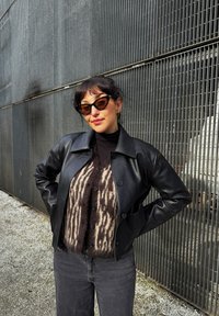 Čierna kožená bunda cez tmavý rolák, doplnená vzorovaným hnedým kardigánom. Doplnky tvoria hranaté slnečné okuliare a kruhové náušnice.