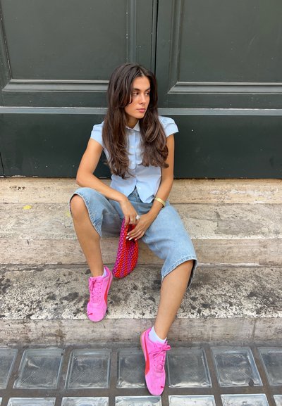 Camisa de manga corta azul claro, pantalones cortos de mezclilla holgados, zapatillas rosas y una bolsa texturizada roja y rosa, sentada en escalones de piedra contra una puerta oscura.
