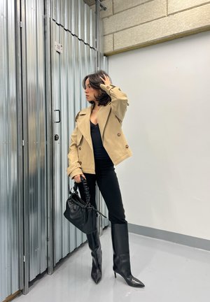 Abrigo trench beige de doble botonadura, top negro ajustado, pantalones pitillo negros, bolso de piel negro, botas de punta altas hasta la rodilla, fondo industrial.