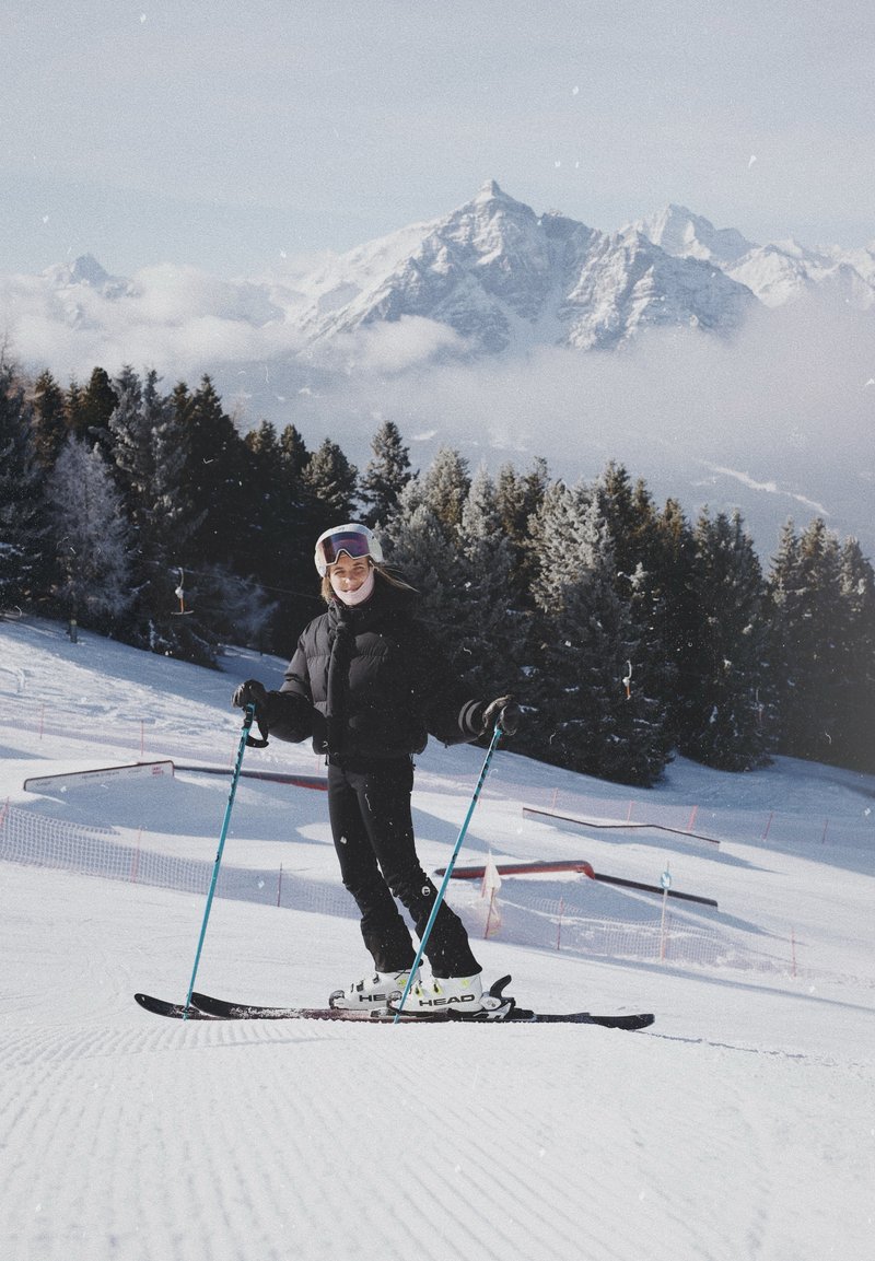 Le matériel de ski comprend des skis noirs et des chaussures de ski blanches. Le skieur porte une veste matelassée noire et un casque de ski violet, sur fond de montagnes enneigées.