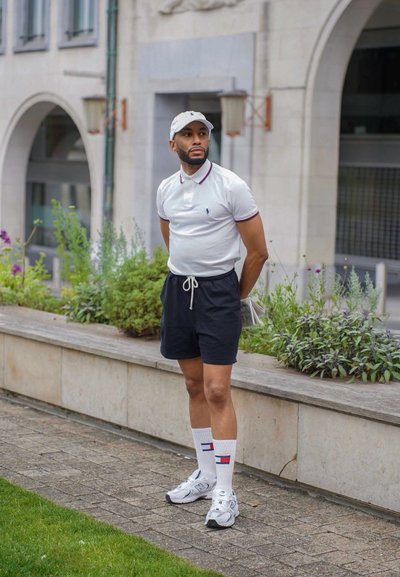 Camisa polo blanca con acentos en rojo y azul marino, pantalones cortos negros, calcetines blancos con rayas rojas y azules, y zapatillas atléticas plateadas. Ambiente casual al aire libre.