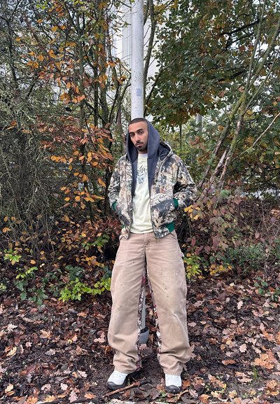 Hombre con chaqueta de camuflaje con capucha y pantalones beige holgados, de pie frente a árboles otoñales y hojas caídas en el suelo.