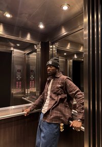 Liftu interjers ar metāliskām sienām, maigu apgaismojumu un elektroniskām pogām. Personas, kas valkā rūtainu brūnu kreklu un zilas džinsa bikses.