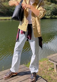 Casaco de canvas bege sobre camisa branca, jeans azul claro com camisa quadriculada atada na cintura, bolsa preta e sapatilhas atléticas castanhas.