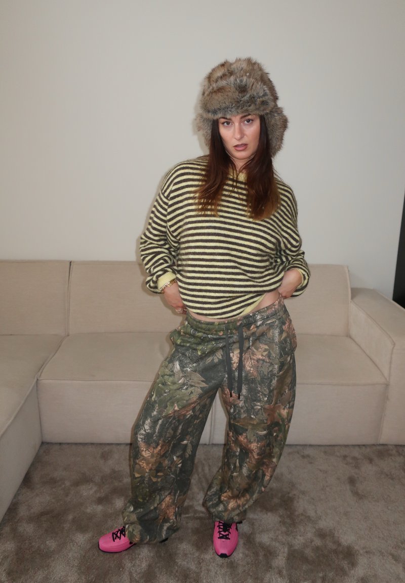 Pulover cu dungi verzi și galbene, pantaloni de camuflaj, adidași roz și o pălărie căptușită cu blană. Stă pe un covor lângă o canapea deschisă la culoare.