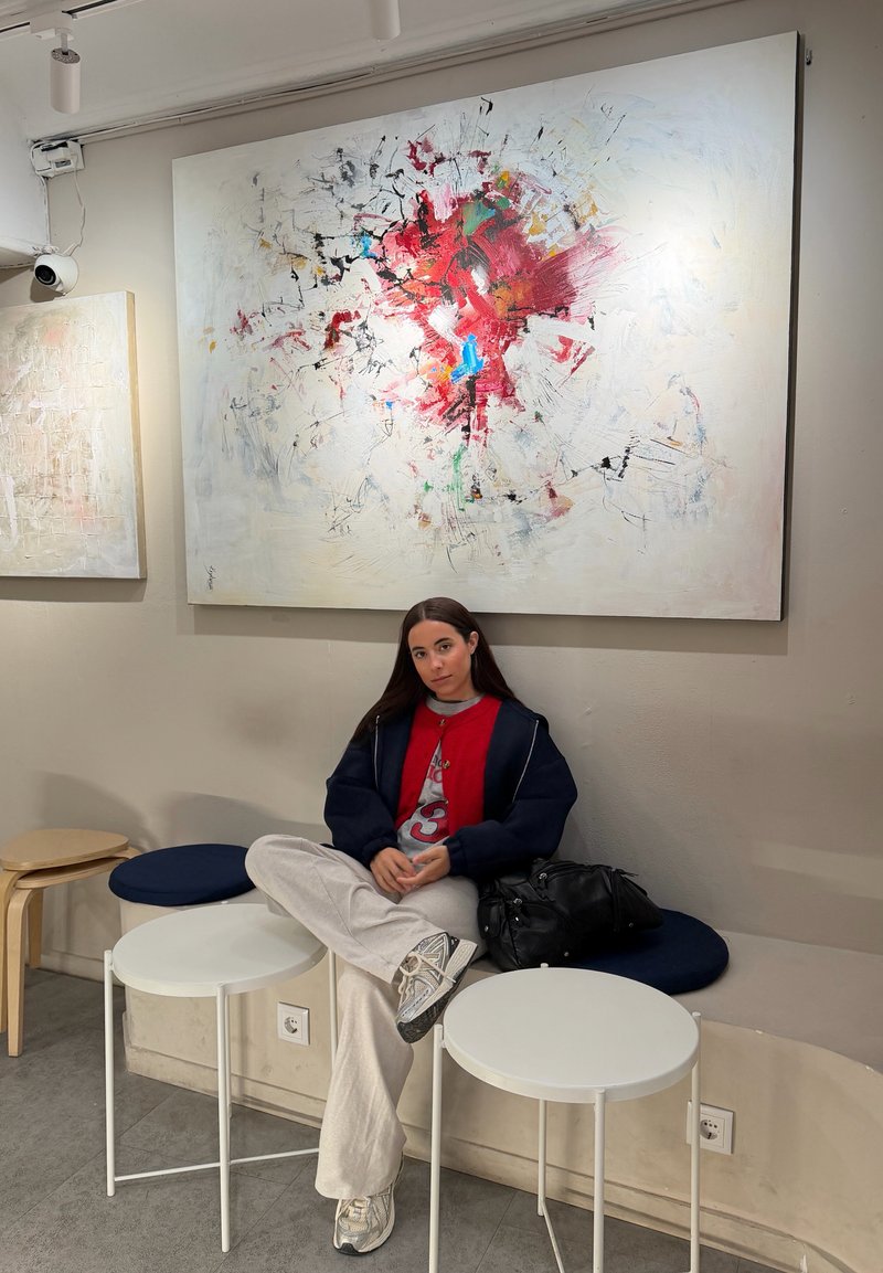 Young woman in casual clothes sitting cross-legged on bench in art gallery under large abstract painting with red and black splashes.