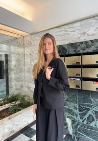 Blazer noir sur mesure avec un bouton unique, associé à un pantalon noir à jambes larges. Tissu lisse, design minimaliste et coupe structurée.