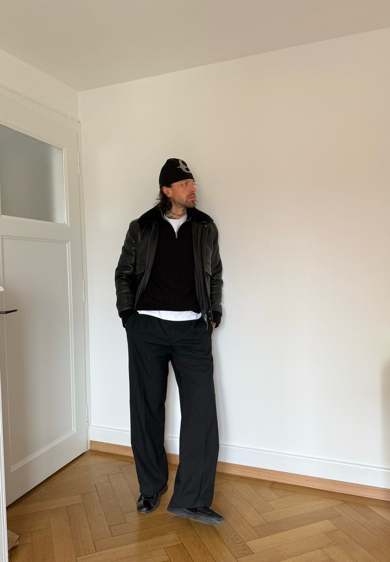 Mężczyzna w czarnej czapce typu beanie, okularach, czarnej skórzanej kurtce, czarnym swetrze, białej koszuli i luźnych czarnych spodniach stoi w pomieszczeniu na drewnianej podłodze.