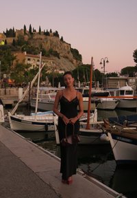 Vestido de noite preto em satin com detalhes de rendas, saltos altos rosa e uma bolsa castanha, de pé ao lado de barcos numa marina ao anoitecer.