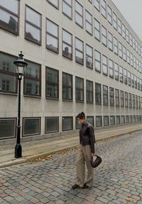 Dunkelbraune, kurze Lederjacke über grauen, weit geschnittenen Hosen, trägt eine braune Handtasche mit Struktur, geht eine Kopfsteinpflasterstraße entlang.