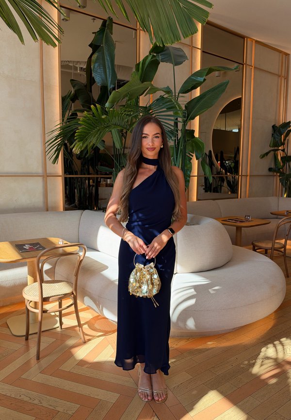 Una mujer con un vestido azul marino sostiene un bolso dorado en un salón luminoso con sofás curvos, mesas de madera y grandes plantas tropicales.