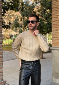 Homme portant un pull beige en maille torsadée, des lunettes de soleil noires et un pantalon en cuir noir texturé, debout à l'extérieur près de colonnes en brique.