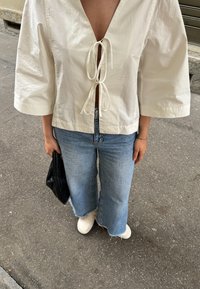 Λευκή, άνετη μπλούζα με βαθύ V-ντεκολτέ και δέσιμο, συνδυασμένη με ανοιχτό μπλε ψιλόλευκες τζιν και κρεμ χρώματος παπούτσια. Περιλαμβάνεται μαύρη τσάντα.