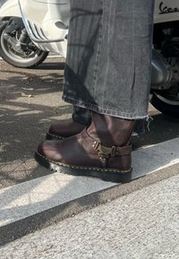 Personne portant des bottes en cuir brun foncé et un pantalon gris, debout sur un passage pour piétons blanc près d'un scooter blanc.