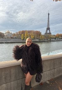 Femme portant un manteau sombre et duveteux ainsi que des bottes, appuyée sur une balustrade en pierre le long de la Seine, avec la tour Eiffel et des arbres d'automne en arrière-plan.