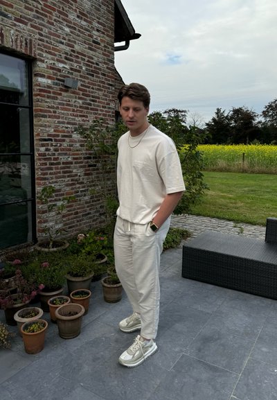 Conjunto informal que incluye una camiseta de algodón beige clara y pantalones de chándal a juego. Zapatillas gris y blancas, reloj delgado, de pie cerca de plantas en macetas.