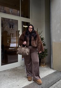 Casaco de pêlo sintético castanho sobre uma blusa preta, calças largas castanhas com riscas laterais, botas estilo ugg castanhas e uma mala cinza texturizada.