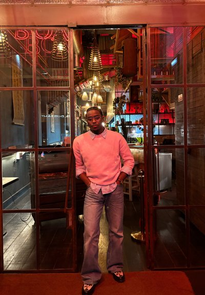 Joven con suéter rosa y vaqueros, de pie en la puerta de un restaurante moderno y cálidamente iluminado con luces colgantes y decoración de madera.