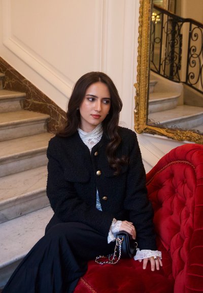 Mujer de cabello oscuro con chaqueta negra y blusa blanca de encaje sentada en un sofá de terciopelo rojo cerca de unas escaleras de mármol y un espejo con marco dorado.