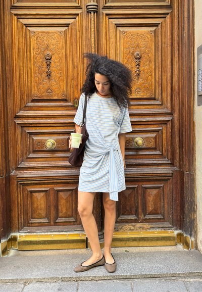 Vestido de camiseta a rayas en azul claro y blanco, con un detalle de lazo en la cintura. Zapatos planos tejidos y un bolso de hombro marrón. Bebida clara en la mano.