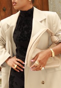 Blazer beige sur une robe noire à motifs floraux, tenant un sac à main tissé de crème. Des bagues en or et une montre mettent en valeur des mains avec des ongles rose pâle.