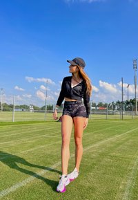 Crna skraćena jakna, crno-sive kratke hlače s uzorkom, bijele i ružičaste sportske tenisice. Zelena travnata površina pod bistrim plavim nebom.