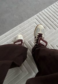 Adidas sporta apavi krēmkrāsā un bordo, ar biezām gumijas zolēm, kontrastējošām svītrām un pārmērīgi lielu mēlīti ar zīmola logotipu. Brūnas bikses uz reljefaina paklāja.