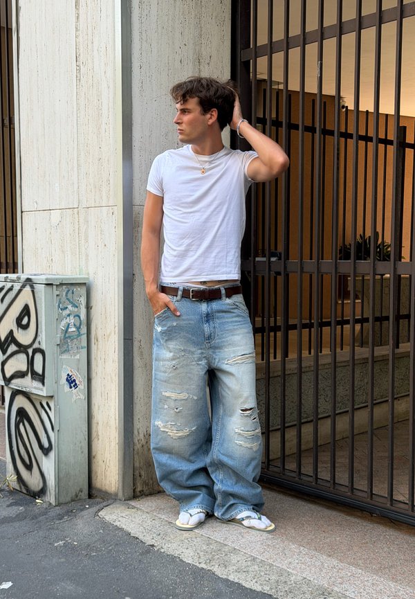 Jeune homme portant un t-shirt blanc et un jean large déchiré, appuyé contre un mur, une main sur la tête et l'autre dans la poche.
