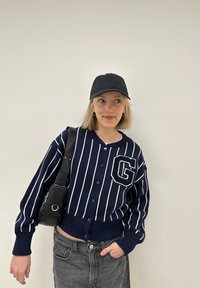 Merimiesraidallinen kardigan etupainikkeilla, jossa on huomattava "G"-patsas. Yhdistetty korkeavyötäröisten harmaiden farkkujen ja mustan lippiksen kanssa.