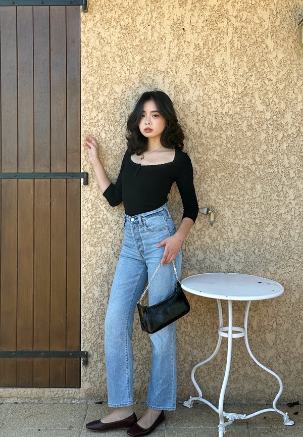 Jeune femme debout contre un mur beige texturé près d'une porte en bois et d'une petite table ronde en métal blanche, portant un haut noir, un jean bleu clair, et tenant un sac à main noir.