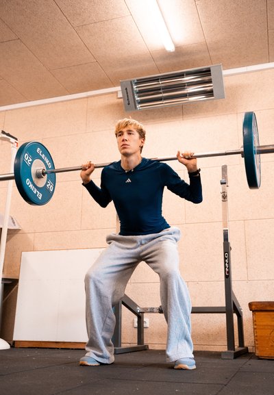 Joven en camiseta azul marino y pantalones de chándal grises levantando una barra con pesas en un gimnasio interior.