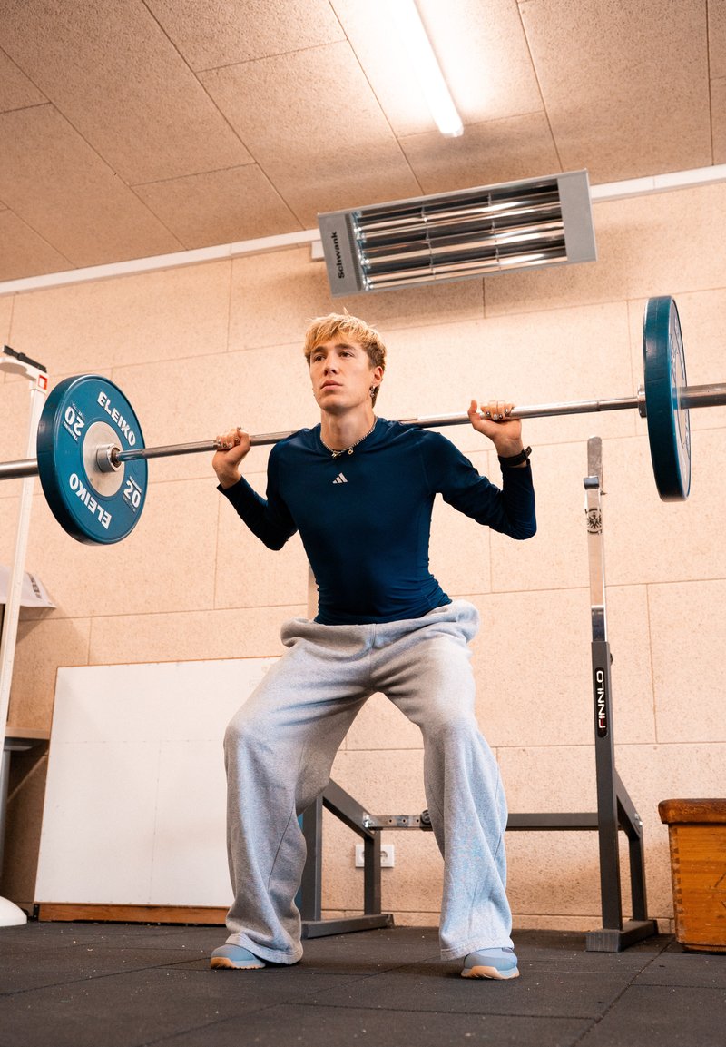Young man in navy shirt and gray sweatpants lifting a barbell with weights in an indoor gym setting.