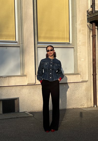 Mujer con cabello rizado que lleva gafas de sol negras, una chaqueta de mezclilla oscura, pantalones negros y zapatos rojos, de pie frente a un edificio beige con persianas amarillas.