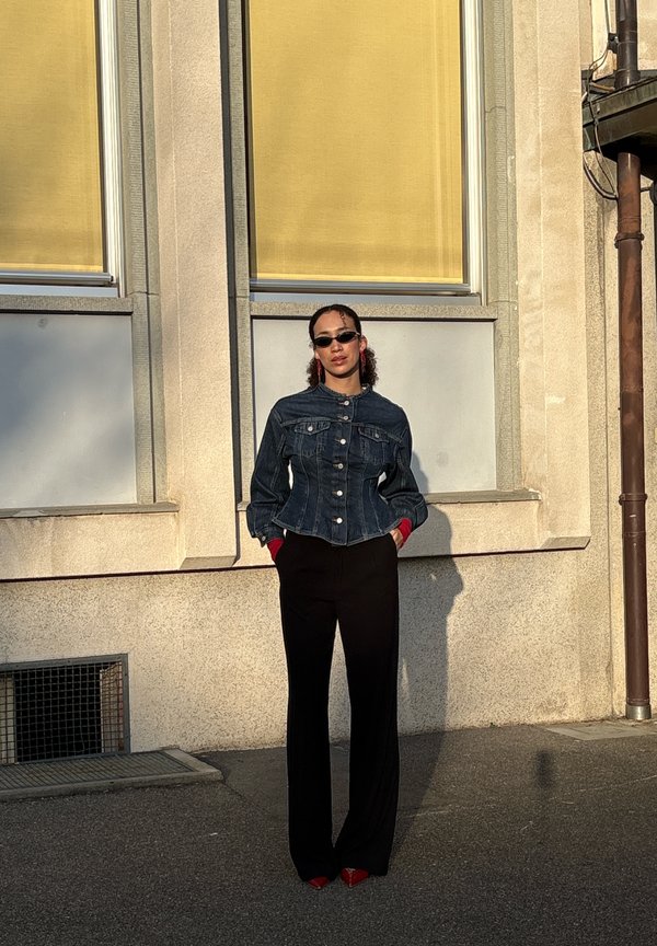 Mujer con cabello rizado que lleva gafas de sol negras, una chaqueta de mezclilla oscura, pantalones negros y zapatos rojos, de pie frente a un edificio beige con persianas amarillas.