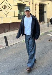 Conjunto casual com uma t-shirt branca, um casaco preto oversized, jeans azuis largos e sapatos castanhos, complementado por um boné camuflado.