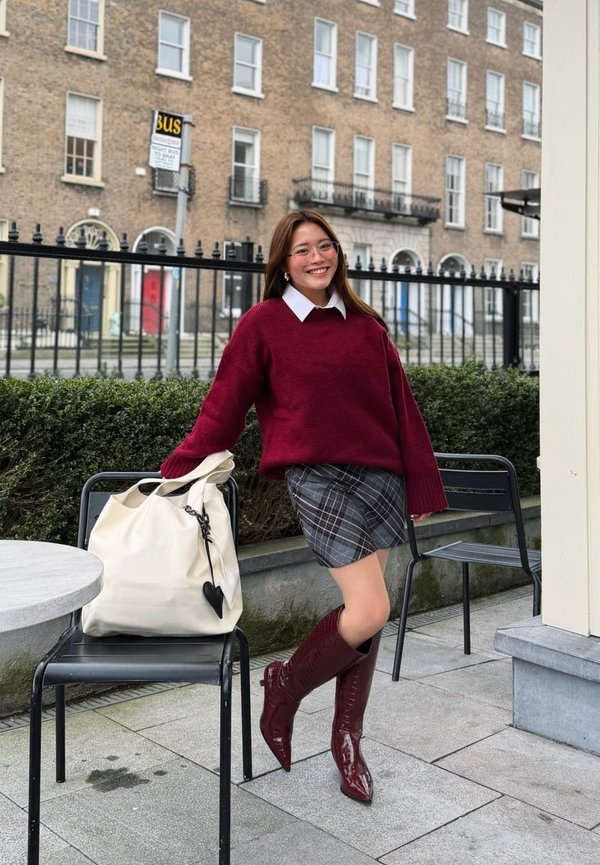 Jeune femme en pull rouge, jupe à carreaux et bottes assorties s'appuie sur une chaise avec un grand sac blanc à l'extérieur, près d'une clôture en fer et d'un bâtiment en briques.