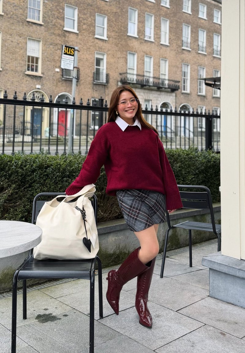 Jeune femme portant un pull rouge, une jupe à carreaux et des bottes assorties, appuyée sur une chaise avec un grand sac blanc à l'extérieur, près d'une grille en fer et d'un bâtiment en briques.