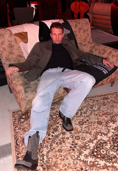 Sofá de tela con estampado floral y patas de madera; individuo con atuendo oscuro, jeans holgados y botas vaqueras descansa con los brazos extendidos sobre la parte trasera.