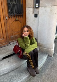 Grønn strikkegenser, brune treningsbukser med gule striper, grå støvler og en rød håndveske. Treport i bakgrunnen med metalletaljer.