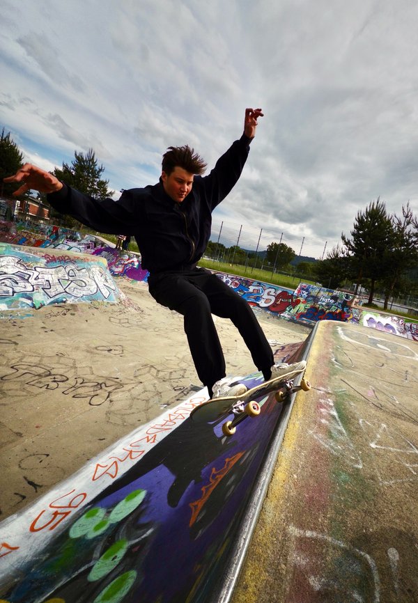 Skateboarder in black clothes performing a grind trick on a graffiti-covered ramp at outdoor skate park under cloudy sky.