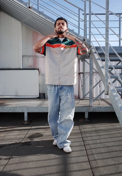 Camisa de manga corta con cuello, con paneles en beige, verde y rojo; etiquetada "KYCAMA". Llevada con jeans desgastados de color azul claro y zapatos blancos.